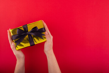 First person top view photo of hands holding golden giftbox with black ribbon bow over shiny golden sequins on isolated red background.