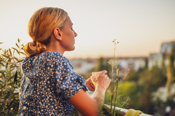 Beautiful woman enjoys drinking coffee while standing on her balcony at sunset.	
