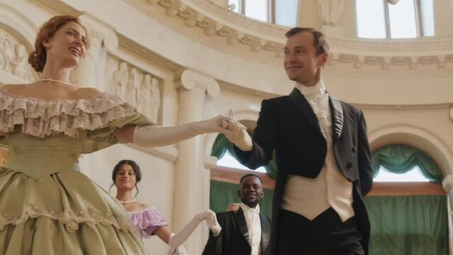 Low angle slowmo of diverse gentlemen wearing elegant tuxedo suits escorting beautiful female debutantes at ball party indoors
