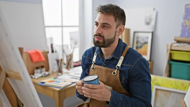 Bearded, young hispanic male artist, doubt in his eyes, nursing his morning coffee cup, deep in thought, looking at a freshly drawn canvas in his bustling art studio.