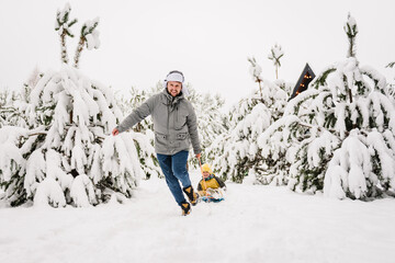 Father pulling little son on winter day. Children are rolling down hill on sledge in forest. Happy funny child with dad ride sled on snowy road in mountains. Family walks during snowfall in park. - obrazy, fototapety, plakaty