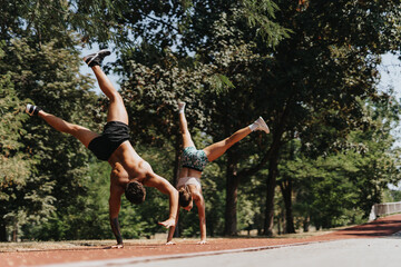 Fit sports duo in a park exudes vitality while practicing outdoor training. They perform confident cartwheels, showcasing athleticism and dedication to better body shape.
