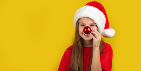 Fototapeta premium Holiday Fun. Cheerful Little Girl Wearing Santa hat, Playful Kid Keeping Xmas Tree Ball Near Nose on Yellow Background. Copy Space