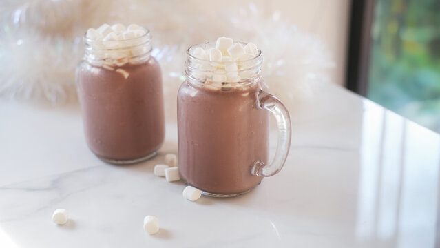 Two transparent mugs with cold cocoa with marshmallows stand on a white kitchen table. There is Christmas tinsel in the background. Christmas summer background image with refreshing drink.
