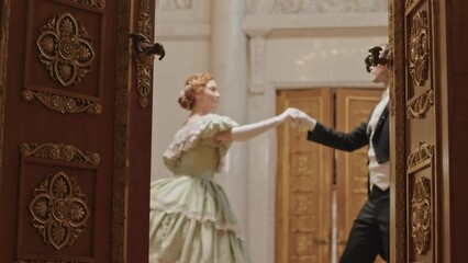 Through wooden door with golden carving details shot of beautiful young Caucasian couple wearing Victorian style outfits dancing on parquet floor in historical ballroom