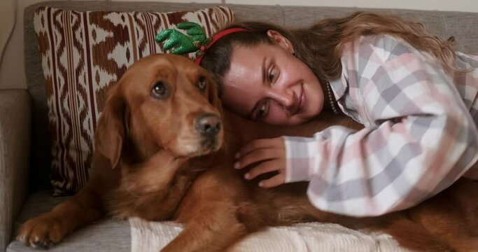 A young woman in a Christmas costume with reindeer antlers lies on the sofa with her golden retriever dog and pets it. Celebrating New Year and Christmas with a dog.