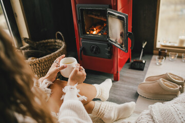 Young woman sitting by the fireplace in white sweater, drinking tea in cozy log cabin.