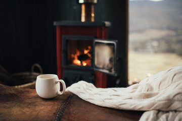 White cup with tea with burning fireplace on the background in cozy log cabin.