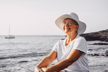 Carefree satisfied senior woman with white hat looking at new day from sea beach at sunrise enjoying freedom, vacation or retirement concept