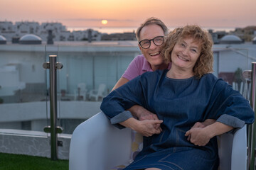 Handsome caucasian mature senior couple smiling sitting on home terrace enjoying sunset over sea looking at camera