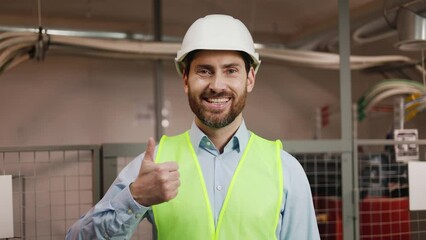 Worker at metallurgical plant, wearing white helmet and vest, stands in electrical switchboard room gives a thumbs up. Engineer technologist electrician smiles at the camera, coordination of actions.