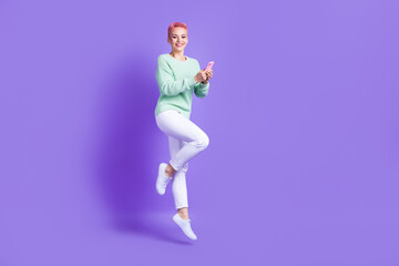 Full length photo of pretty cute girl wear sweater texting apple samsung device jumping high empty space isolated violet color background
