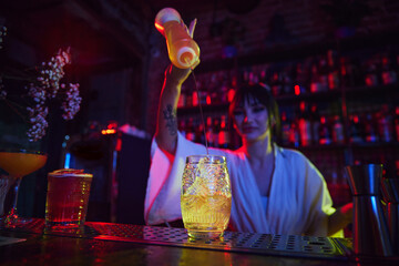 Beautiful young girl working as bartender in modern bar, making delicious cocktail, adding liquids into glass with ice. Concept of occupation, nightlife, bar, party, alcohol drink, mixologist