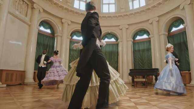 Full length slowmo of three multiracial aristocratic couples in fancy Victorian outfits dancing energetically on parquet floor in posh ballroom