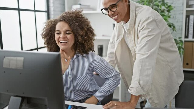 Two indoor office workers, a man and a woman, confidently raise up their hands for a high five, expressing professional success at their workplace computer desk.