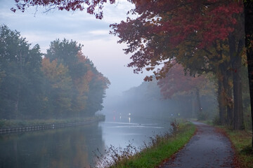 The image captures a serene canal scene, enveloped in the soft mist of an autumn morning. The...