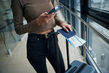 Holding smartphone. Close up view of female traveler in airport hall with tickets - obrazy, fototapety, plakaty