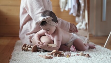 A baby girl lies on her stomach on her mother lap and curiously studies environmental toys while...