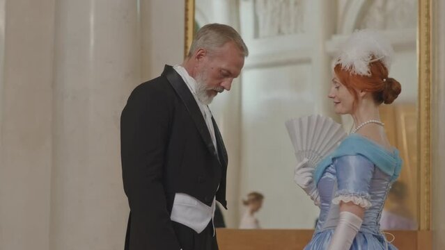 Medium slowmo of good looking red haired Caucasian woman with fan in hands speaking to male guest at beautiful ceremonial reception in spacious classical ballroom