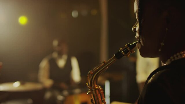 Side view medium closeup of young Black female saxophonist playing instrument while performing on stage with jazz band on stage in fancy club
