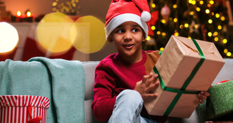 Obraz na płótnie Canvas Close up portrait of cute small African American boy in santa hat sitting at decorated home in evening on New Year's Eve with excited face holding xmas gift in hands with glowing tree on background