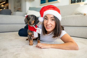 Young smiling woman with her cute dachshund dog wearing santa claus hat at home.