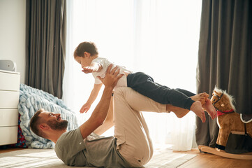 Father is lying down, holding boy in hands. Man is playing with his little son in the domestic room