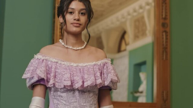 Tilting up slowmo portrait of young Biracial aristocratic girl wearing poof ruffled purple ball dress posing for camera in classic ball hall