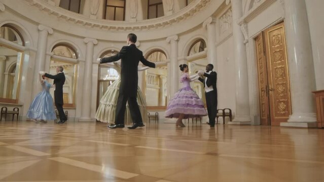 Slowmo long shot of beautiful women in poof dresses and their male companions ball dancing on parquet floor in spacious classic ballroom with ornamental walls and windows