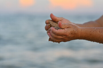 Close-up elderly man's hands releasing and dropping sand. Sand flows through the hands against the blue ocean on the background. Summer beach holiday vacation concept
