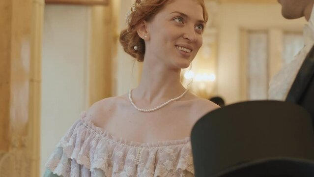 Tilting up shot of young beautiful Caucasian woman wearing elegant ball dress and long gloves having conversation with male companion during ceremonial reception in luxury mansion