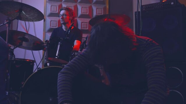 A singer is emotionally singing a song into a microphone, dramatically sitting on the floor in a music studio next to speakers and a drum kit on which a drummer is playing.
