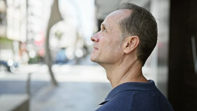 Middle age man, relaxed yet with a serious expression, standing outside looking up to the city's sky from a bustling urban street