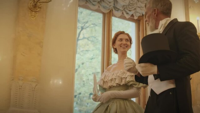 Low angle medium slowmo of Caucasian young woman in beautiful ball dress listening to mid aged male companion while strolling together along classic ball hall during gala reception