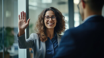 Cheerful businesswoman wearing eyeglasses and a business suit, raising her hand in a greeting an office meeting, with colleagues - obrazy, fototapety, plakaty