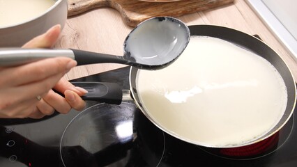Pouring Batter onto Pan: A person's hand holds a ladle, guiding a smooth flow of batter onto a hot pan. Adjacent to this, a cooked crepe rests on a wooden board. Making pancake. Cooking food.  - obrazy, fototapety, plakaty