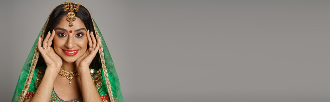 joyful indian woman with bindi dot and green veil smiling at camera with hands near face, banner