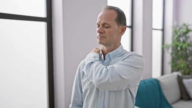 Hispanic middle age man, looking cool yet serious, suffering from intense shoulder pain indoors - an adult, stressed from his injury, standing strong in his living room