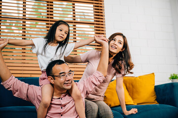 A happy Asian family father mother and daughter enjoy quality time on a comfortable sofa in their modern house. Togetherness laughter and bonding during self-isolation bring joy.
