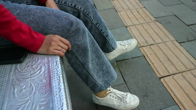 close up of legs of a woman wearing white shoes and jeans sitting in a city park swinging her legs. Relax waiting for someone