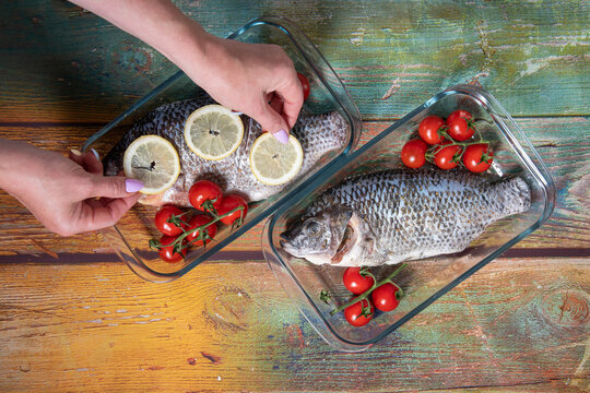 female hand puts lemon slices on fresh tasty raw fish tilapia or dorado before baking in the oven with vegetables and spices, on old green wood, top view, high quality photo