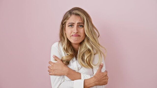 Heartbroken young blonde woman in shirt shivers, sad expression on her face from winter's cold shock, standing alone against pink background
