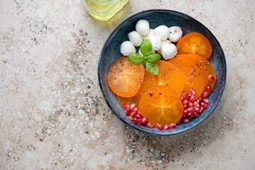 Blue bowl with sliced persimmon, mozzarella balls and pomegranate, horizontal shot on a beige...