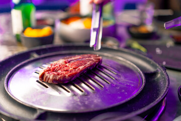 close-up of a hand with tongs picking up a juicy raw piece of meat to put on the grill in a Korean restaurant - obrazy, fototapety, plakaty