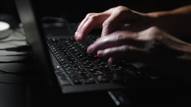 footage of a man's hands typing on a laptop keyboard 
dark lightning room in the frame, black colored laptop, light falls on hands