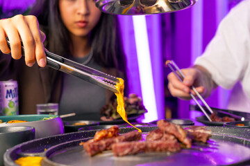 close-up guests of a Korean restaurant take fried meat from the grill with chopsticks and dip it in...