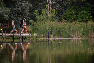 Fit girls warm up together, stretching on a bridge in a green park surrounded by nature and water. Their athletic bodies reflect a dedication to a healthy and active lifestyle.