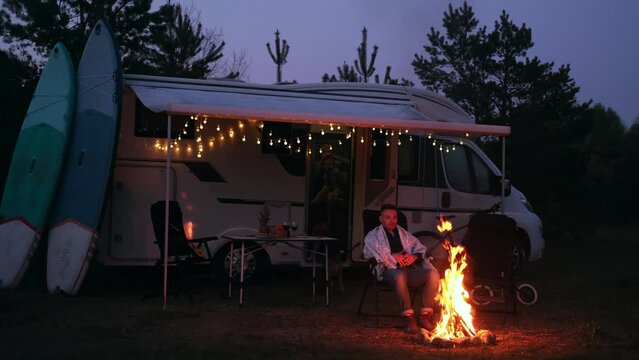 A man is sitting by the campfire in the evening, wrapped in a blanket. Evening rest near a mobile home