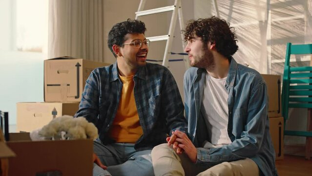 Portrait of a happy multiethnic gay couple smiling looking at camera, in a new house, full of boxes.
The gay couple is happy, they hug and smile.