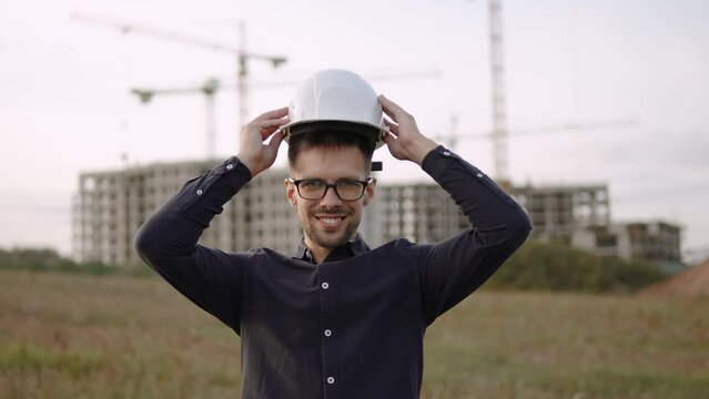 Caucasian male builder engineer with glasses puts on helmet with smile. Compliance with construction site safety regulations during presentation. Portrait of architect caring about health at work.